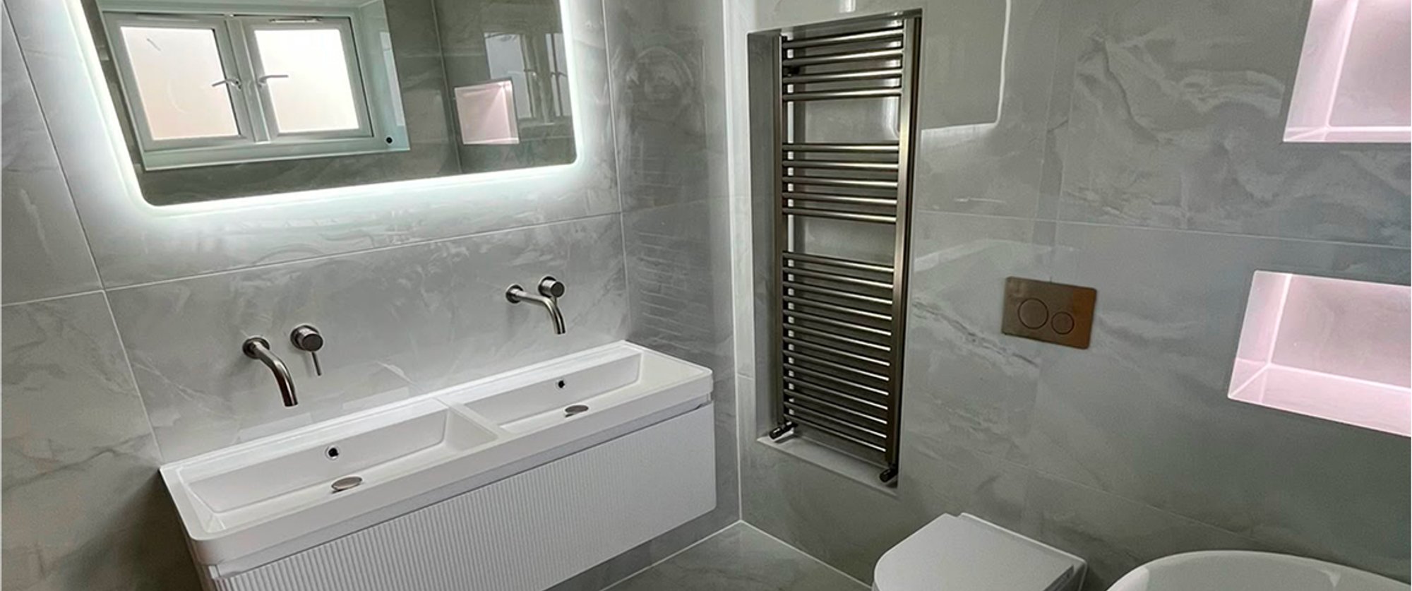 Modern grey bathroom with an illuminated mirror, twin basins, towel radiator, toilet and bath